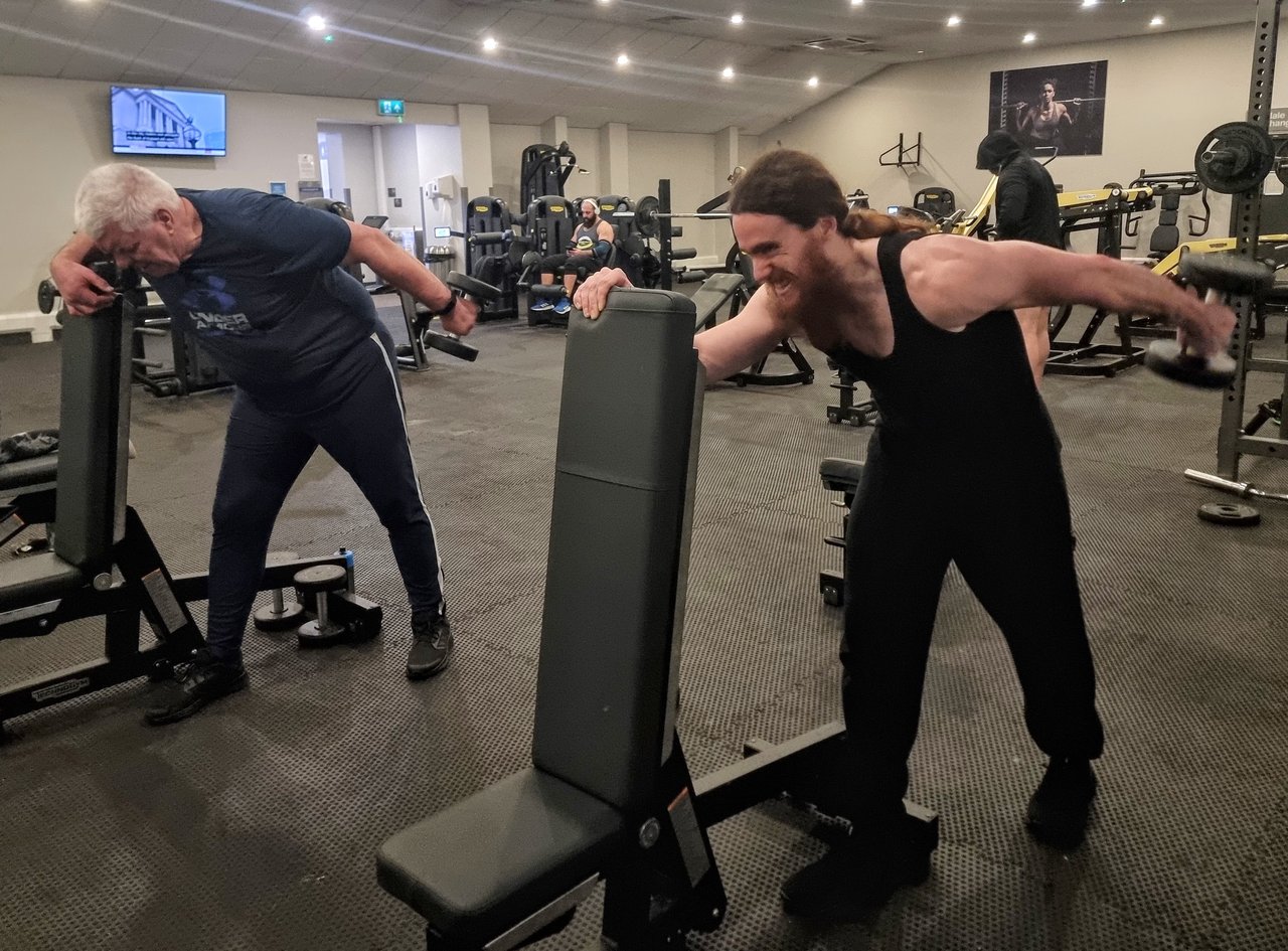 A personal trainer and his client both performing a shoulder exercise in a gym, displaying exertion